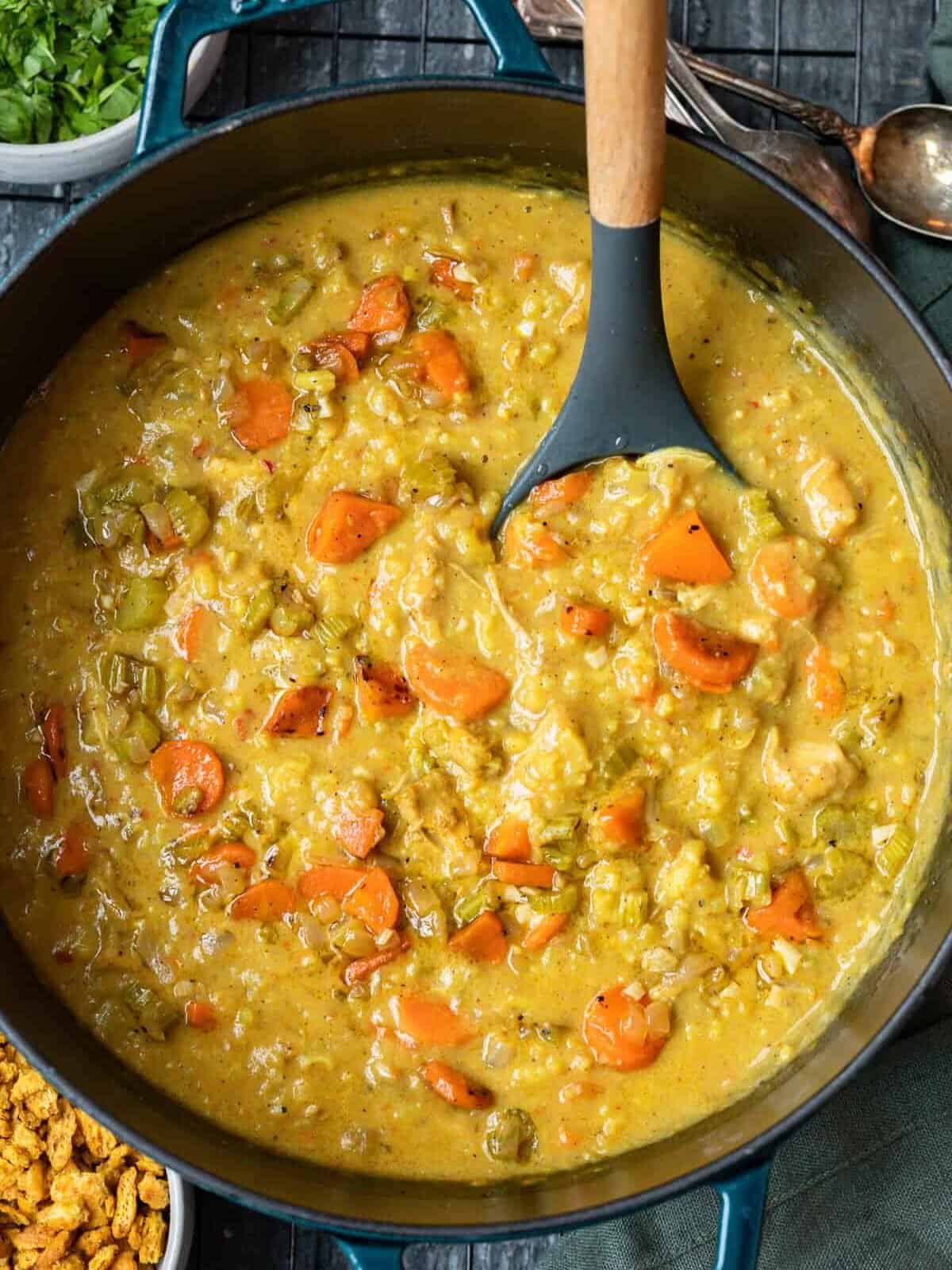 vista aérea da sopa mulligatawny indiana em um forno holandês com uma colher de servir. Cenouras fatiadas, aipo, frango e arroz são visíveis na base cremosa de curry.