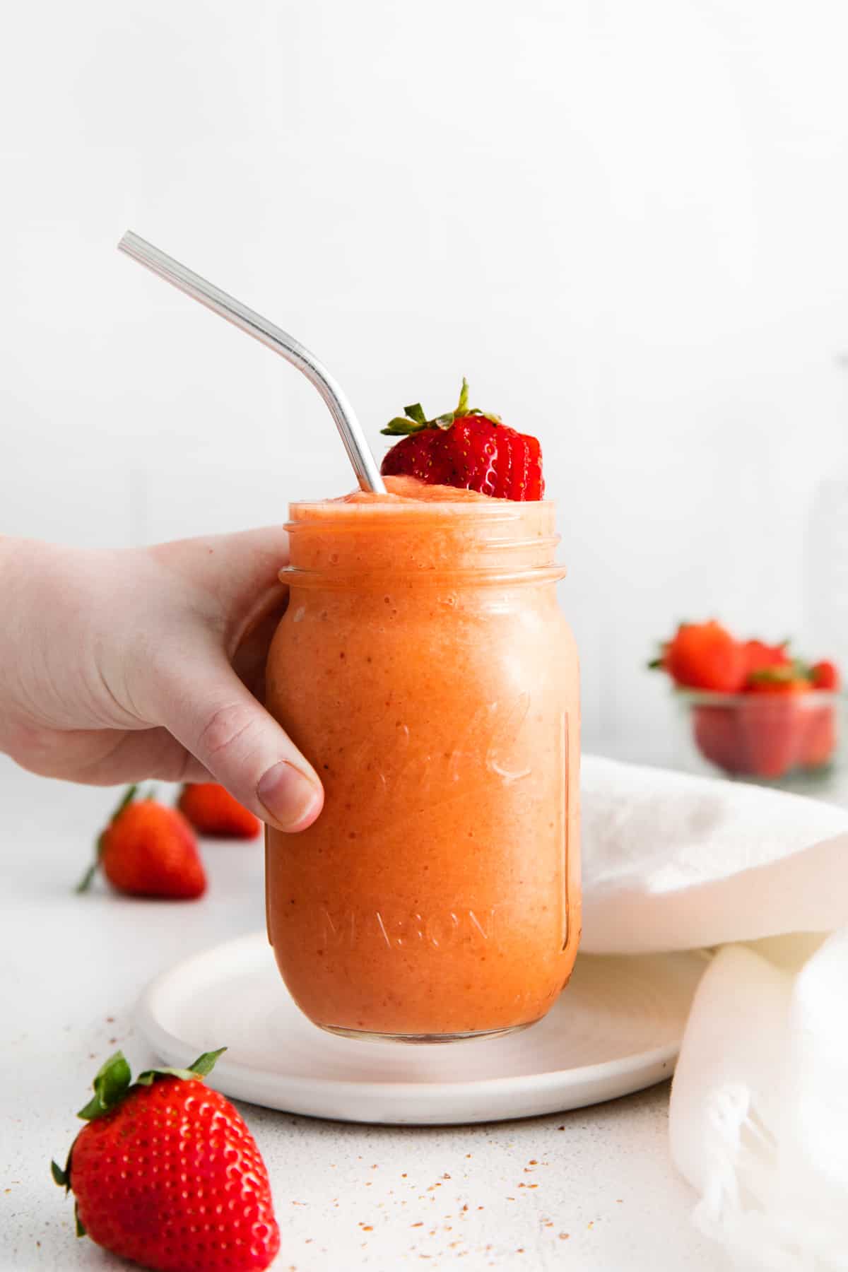 a hand grabbing a strawberry mango smoothie in a mason jar with a stainless steel straw.