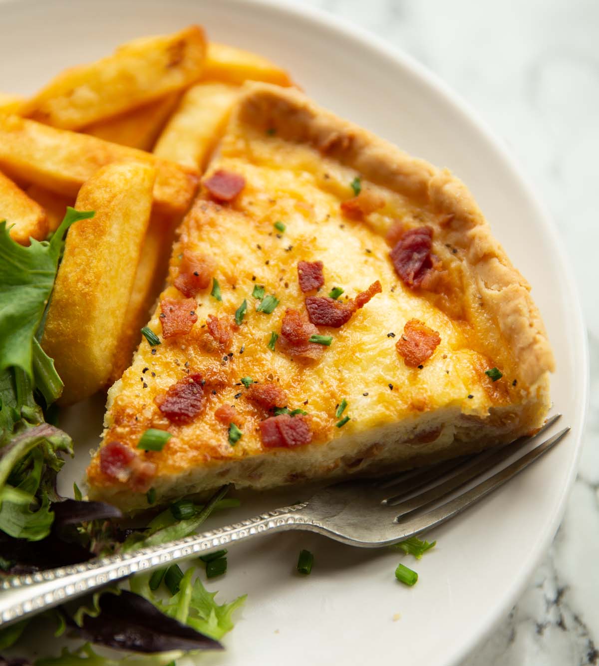 close up shot of slice of quiche lorraine on small white plate with chips and salad