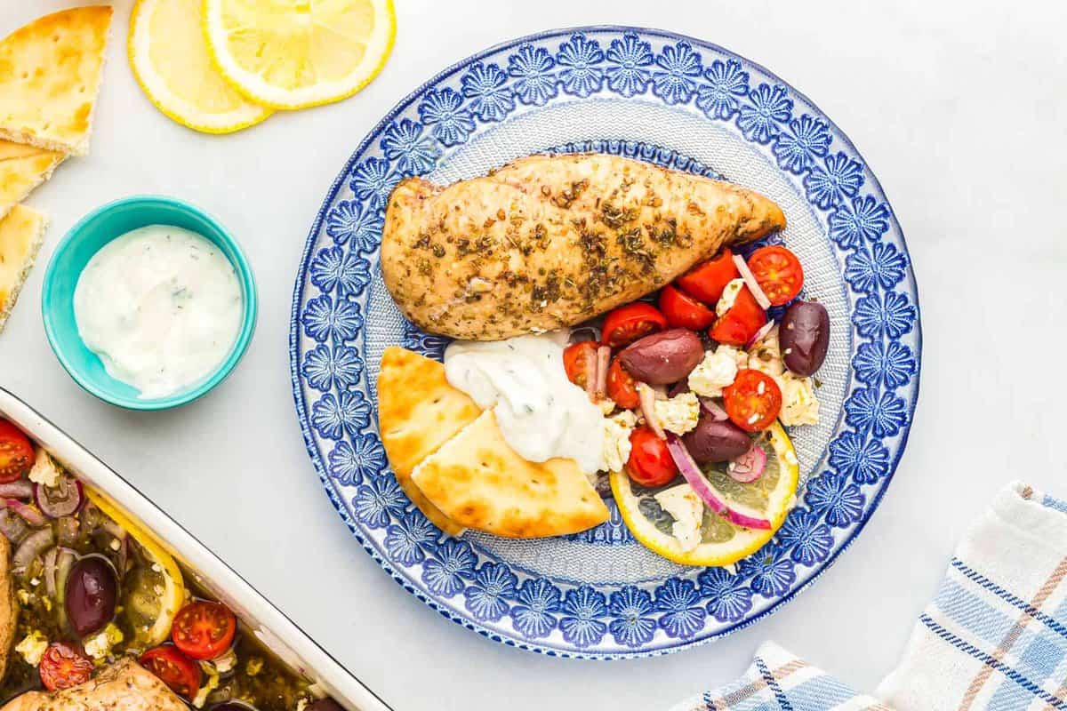 visão aérea de um peito de frango grego assado em um prato estampado azul com pão pita e iogurte.